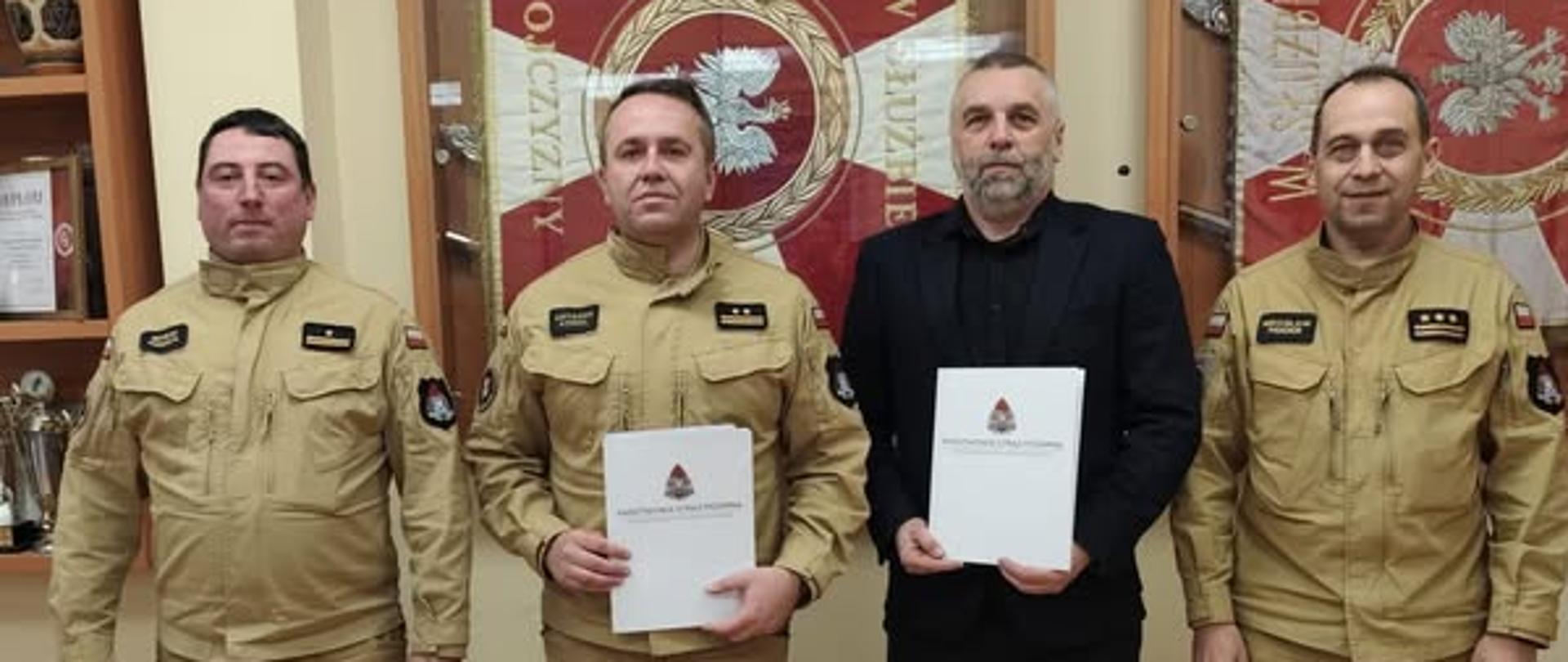 Na zdjęciu są czterej mężczyźni.
Trzech mężczyzn nosi jasne mundury strażackie.
Jeden mężczyzna, drugi z prawej strony, jest ubrany w ciemny garnitur.
Dwóch mężczyzn trzyma białe teczki z logo Państwowej Straży Pożarnej.
Wszyscy stoją obok siebie i patrzą w stronę aparatu.
Za nimi widać strażackie sztandary w kolorze czerwonym oraz półkę z pucharami.