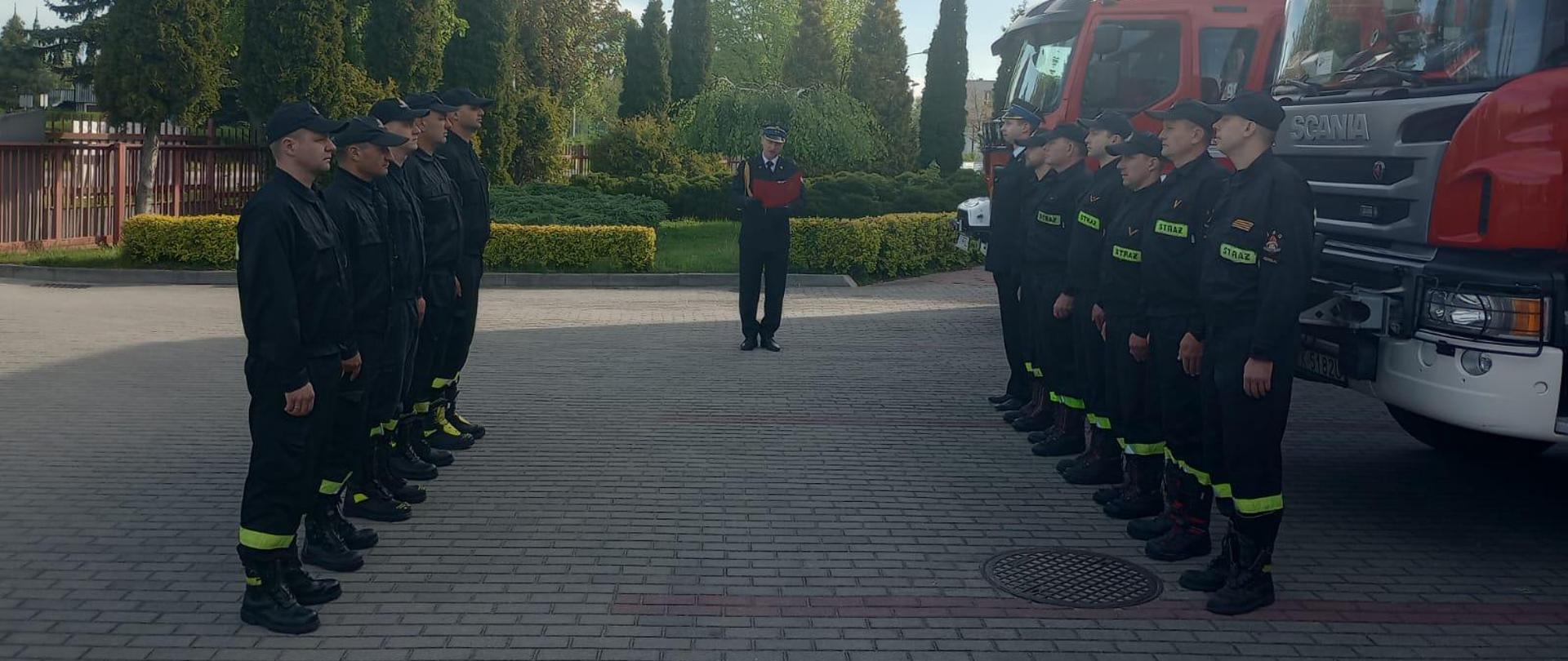 Uroczysta zbiórka przed pojazdami pożarniczymi. Strażacy stoją w dwóch rzędach na przeciwko siebie. Zastępca komendanta powiatowego przedstawia tło historyczne Dnia Flagi Rzeczypospolitej Polskiej.