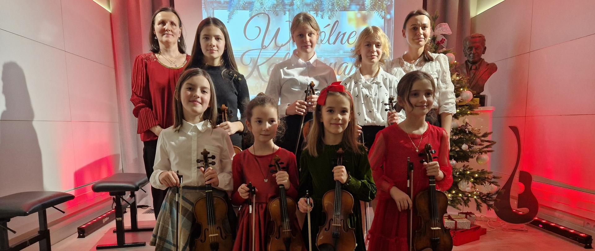 Zdjęcie grupowe klasy skrzypiec na stopniach sceny w dwurzędzie. Uczennice odświętnie ubrane trzymają w dłoniach skrzypce. Z prawej strony stoi nauczycielka, a z lewej akompaniatorka. W tle wyświetlony jest slajd z napisem "Wspólne Kolędowanie". Całość dopełnia oświetlenie w kolorze czerwonym. 