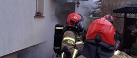 Na zdjęciu strażacy PSP w Grajewie w ubraniach specjalnych i z aparatami oddechowymi na plecach przed wejściem do budynku mieszkalnego jednorodzinnego, z którego wydobywa się dym. W tle budynek mieszkalny jednorodzinny z otwartymi drzwiami wejściowymi, z których wydobywa się dym.