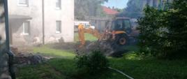 Zdjęcie przedstawia koparkę stojącą przed budynkiem, która przerwała rurę gazową. Przed koparka znajduje się kurtyna wodna w celu zabezpieczenia wycieku gazu.