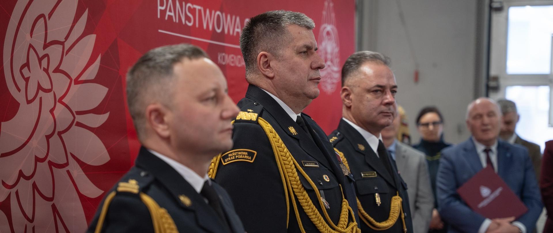 Zdjęcie przedstawia trzech oficerów straży pożarnej stojących w rzędzie w półprofilu. Mają na sobie ciemnogranatowe mundury wyjściowe ze złotymi sznurami galowymi. W tle widać fragment czerwonej ścianki promocyjnej z białym orłem. Po prawej stronie, w głębi, stoją dwie osoby cywilne, z których jedna trzyma czerwoną teczkę z dokumentami.