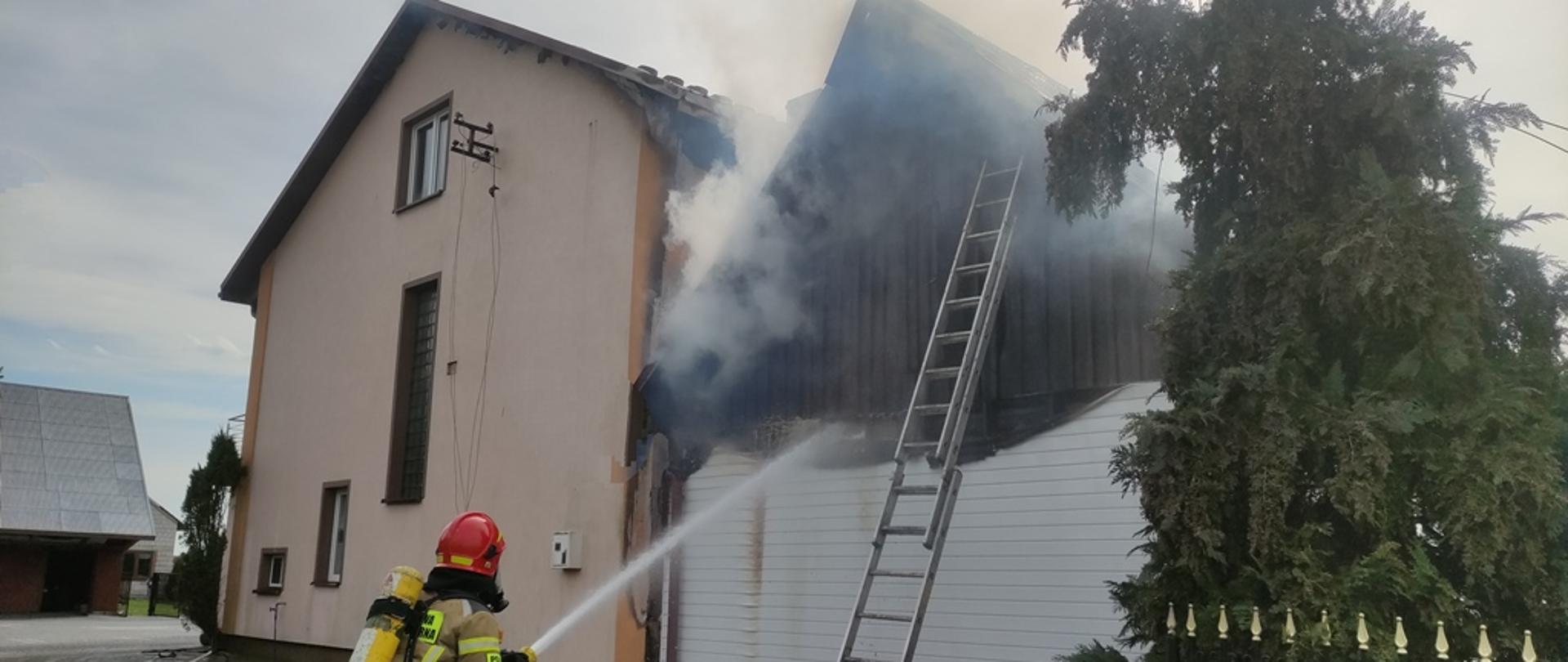 Dwa budynki mieszkalne w bliskiej odległości od siebie, dym wydobywający się spod pokrycia dachowego drewnianego, strażak w sprzęcie ochrony układu oddechowego podje prąd wody na pożar