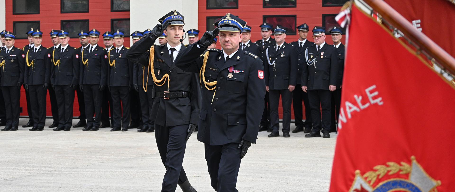 Zastępca Mazowieckiego Komendanta Wojewódzkiego PSP st. bryg. Maciej Bieńczyk i dowódca uroczystości oddają honor przed sztandarem widocznym na pierwszym planie. W tle strażacy PSP i OSP.