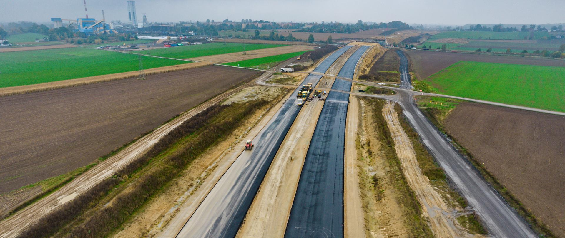 Postępy prac na drodze ekspresowej S1 z lotu ptaka. Trwa budowa drogi w nowym śladzie. Widoczne asfaltowanie. Przed nami powstaje wiadukt WS-11, który przeprowadzi S1 nad linią kolejową w Bieruniu. Widok trasy w kierunku Bielska-Białej.