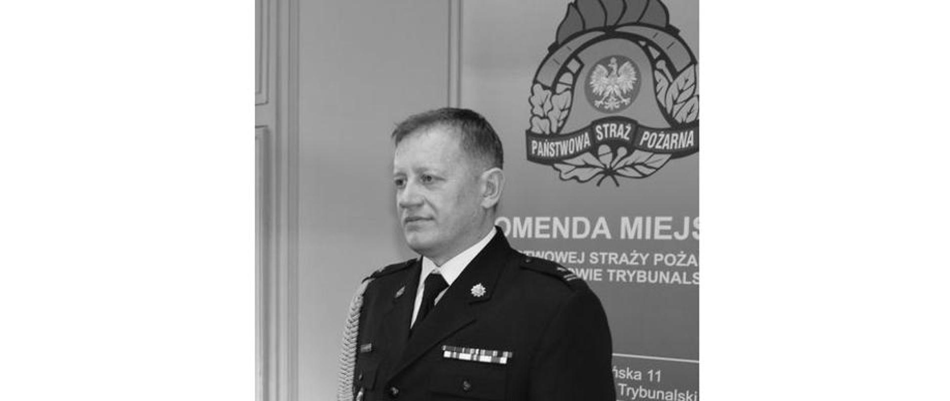 czarno białe zdjęcie przedstawiające Macieja Dobrakowskiego w mundurze wyjściowym za nim rollup komendy miejskiej PSP w Piotrkowie Trybunalskim