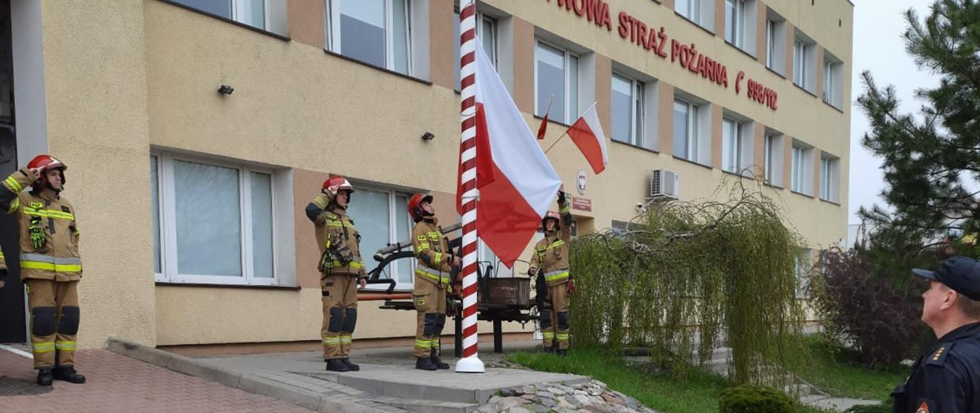 Strażacy ustawieni w szeregu przed budynkiem komendy, przed biało-czerwonym masztem z wciągniętą flagą Polski na maszt. Po lewej strażacy ubrani w piaskowe ubranie specjalne oraz czerwony hełm. Po prawej w czarnym koszarze dowódczo sztabowym.
