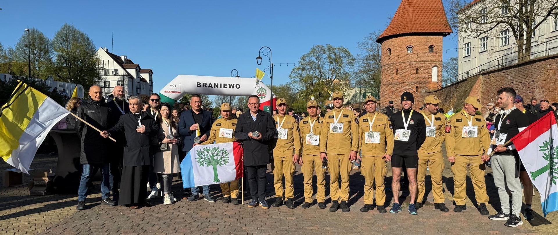 Grupa zawodników, strażaków w jasnych mundurach oraz organizatorzy biegu. Słonecznie.