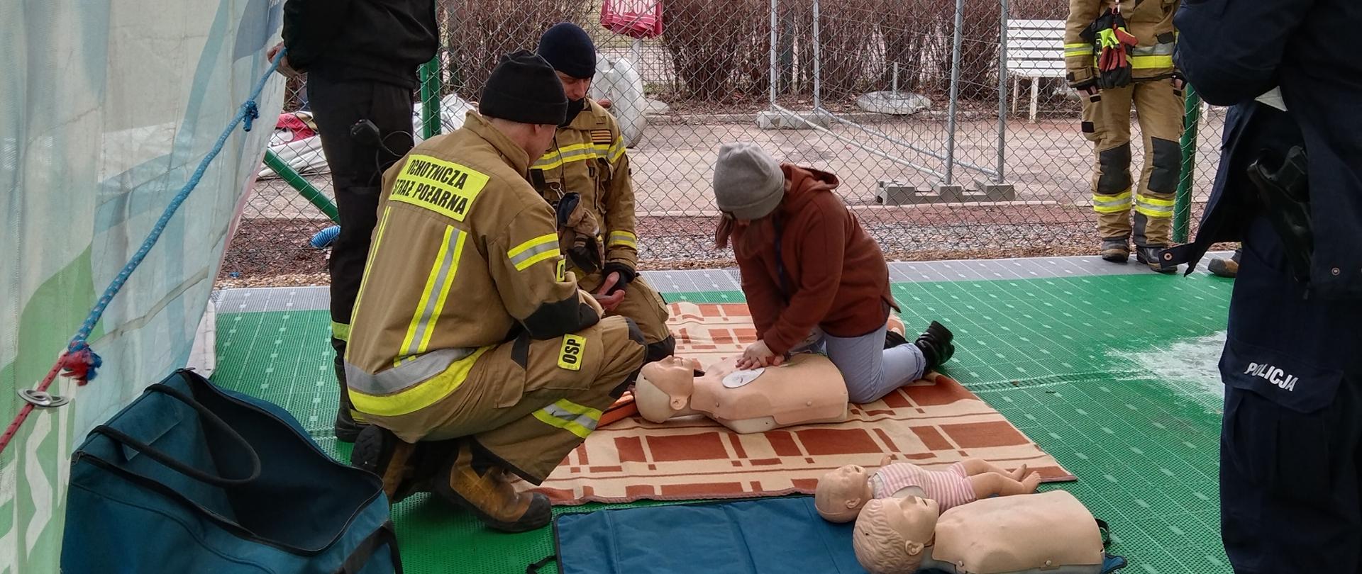 Zdjęcie przedstawia strażaków szkolących dziecko z zakresu pierwszej pomocy.