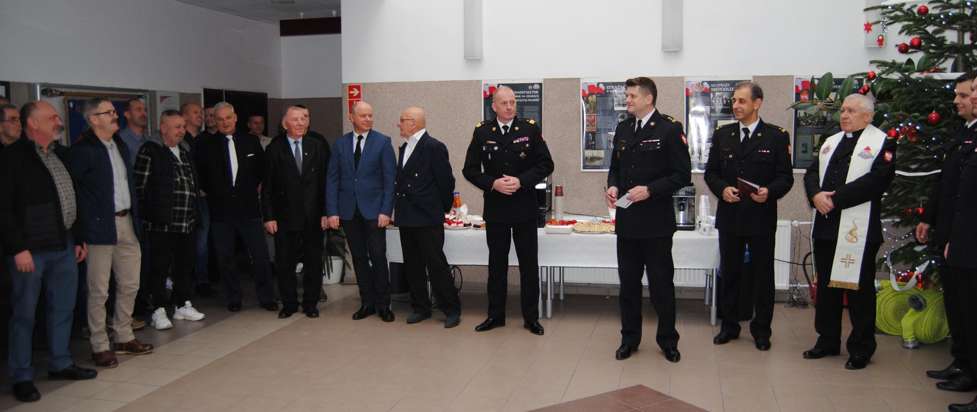 w pomieszczaniu zebrali się ludzie po lewej grupa emerytów następnie prezes emerytów, dwaj byli komendanci, komendant wojewódzki, komendant powiatowy oraz zastępca, kapelan wojewódzki. Po prawej choinka przystrojona czerwonymi bombkami.
