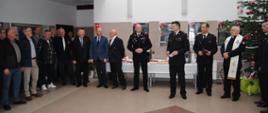 w pomieszczaniu zebrali się ludzie po lewej grupa emerytów następnie prezes emerytów, dwaj byli komendanci, komendant wojewódzki, komendant powiatowy oraz zastępca, kapelan wojewódzki. Po prawej choinka przystrojona czerwonymi bombkami.