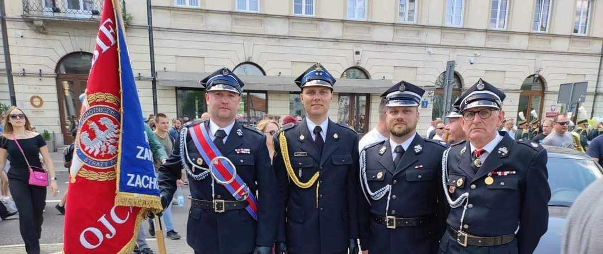 Centralny punkt zdjęcia stanowi czterech strażaków w mundurach wyjściowych. Pierwszy od lewej trzyma sztandar. Drugi jest w mundurze wyjściowym PSP a trzej pozostali OSP. W tle kamienica.