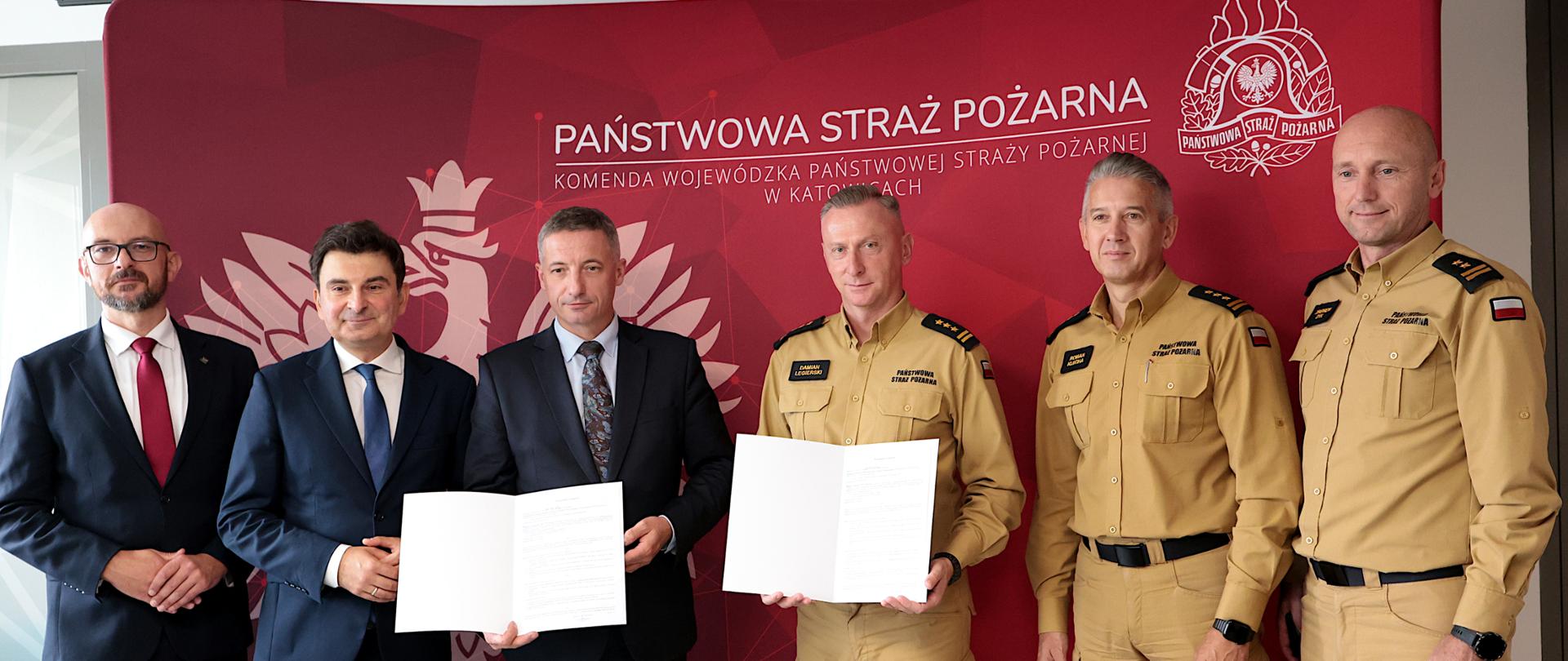 Na zdjęciu sześć osób (mężczyzn) stoi przed czerwonym tłem z logo i nazwą Komendy Wojewódzkiej Państwowej Straży Pożarnej w Katowicach. Na jej środku umieszczono biały napis „Państwowa Straż Pożarna”, a poniżej mniejszy tekst „Komenda Wojewódzka Państwowej Straży Pożarnej w Katowicach”. Po prawej stronie widoczne jest duże godło Państwowej Straży Pożarnej w kolorze białym.
Trzech mężczyzn ubranych jest w ciemne garnitury są to Przewodniczący Śląskiego Związku Gmin i Powiatów - Pan Piotr Kuczera oraz dyrektor biura Związku - Pan Witold Magryś oraz wojewoda śląski - Pan Marek Wójcik. Od prawej trzech strażaków w piaskowych mundurach służbowych - śląski komendant wojewódzki Państwowej Straży Pożarnej - st. bryg. Damian Legierski oraz zastępcy śląskiego komendanta wojewódzkiego st. bryg. Roman Klecha i bryg. Zbigniew Dyk.
Dwaj uczestnicy trzymają otwarte teczki z treścią podpisanego porozumieniem. Wszyscy stoją obok siebie, zwróceni w stronę obiektywu i uśmiechają się do zdjęcia.