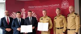 Na zdjęciu sześć osób (mężczyzn) stoi przed czerwonym tłem z logo i nazwą Komendy Wojewódzkiej Państwowej Straży Pożarnej w Katowicach. Na jej środku umieszczono biały napis „Państwowa Straż Pożarna”, a poniżej mniejszy tekst „Komenda Wojewódzka Państwowej Straży Pożarnej w Katowicach”. Po prawej stronie widoczne jest duże godło Państwowej Straży Pożarnej w kolorze białym.
Trzech mężczyzn ubranych jest w ciemne garnitury są to Przewodniczący Śląskiego Związku Gmin i Powiatów - Pan Piotr Kuczera oraz dyrektor biura Związku - Pan Witold Magryś oraz wojewoda śląski - Pan Marek Wójcik. Od prawej trzech strażaków w piaskowych mundurach służbowych - śląski komendant wojewódzki Państwowej Straży Pożarnej - st. bryg. Damian Legierski oraz zastępcy śląskiego komendanta wojewódzkiego st. bryg. Roman Klecha i bryg. Zbigniew Dyk.
Dwaj uczestnicy trzymają otwarte teczki z treścią podpisanego porozumieniem. Wszyscy stoją obok siebie, zwróceni w stronę obiektywu i uśmiechają się do zdjęcia.