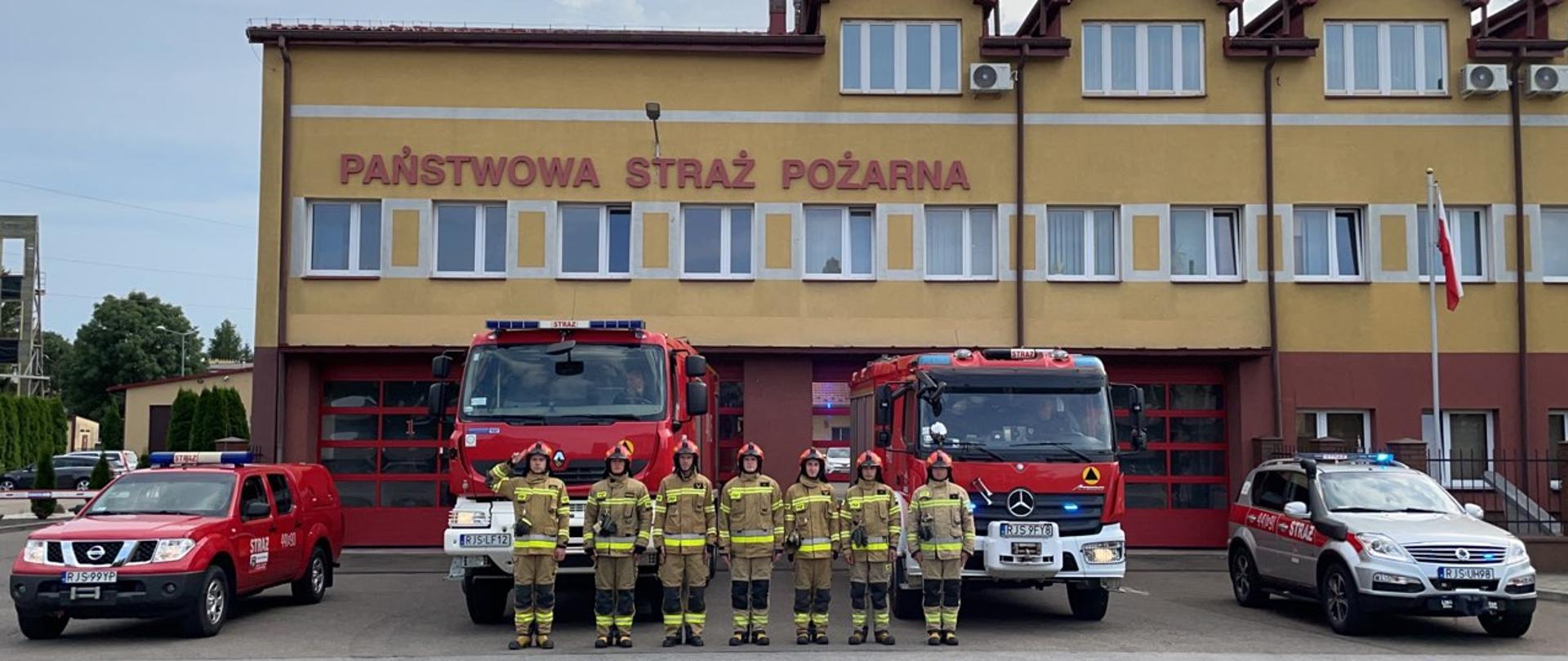 na zdjęciu zmiana służbowa jednostki ratowniczo gaśniczej w Jaśle w umundurowaniu na tle komendy, wozy strażackie przed jednostką