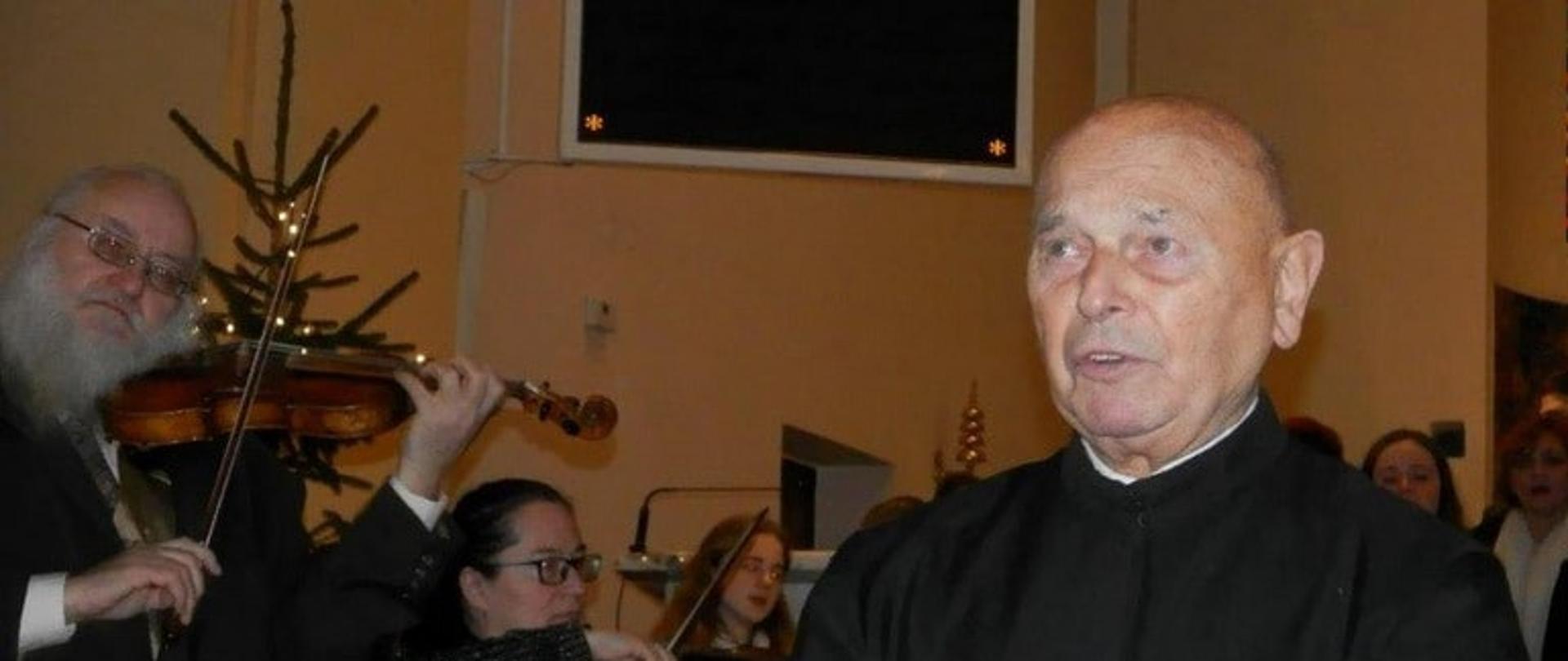 Zdjęcie przedstawiające księdza Wilczyńskiego w sutannie, w trakcie koncertu, w tle muzycy grający na skrzypcach.