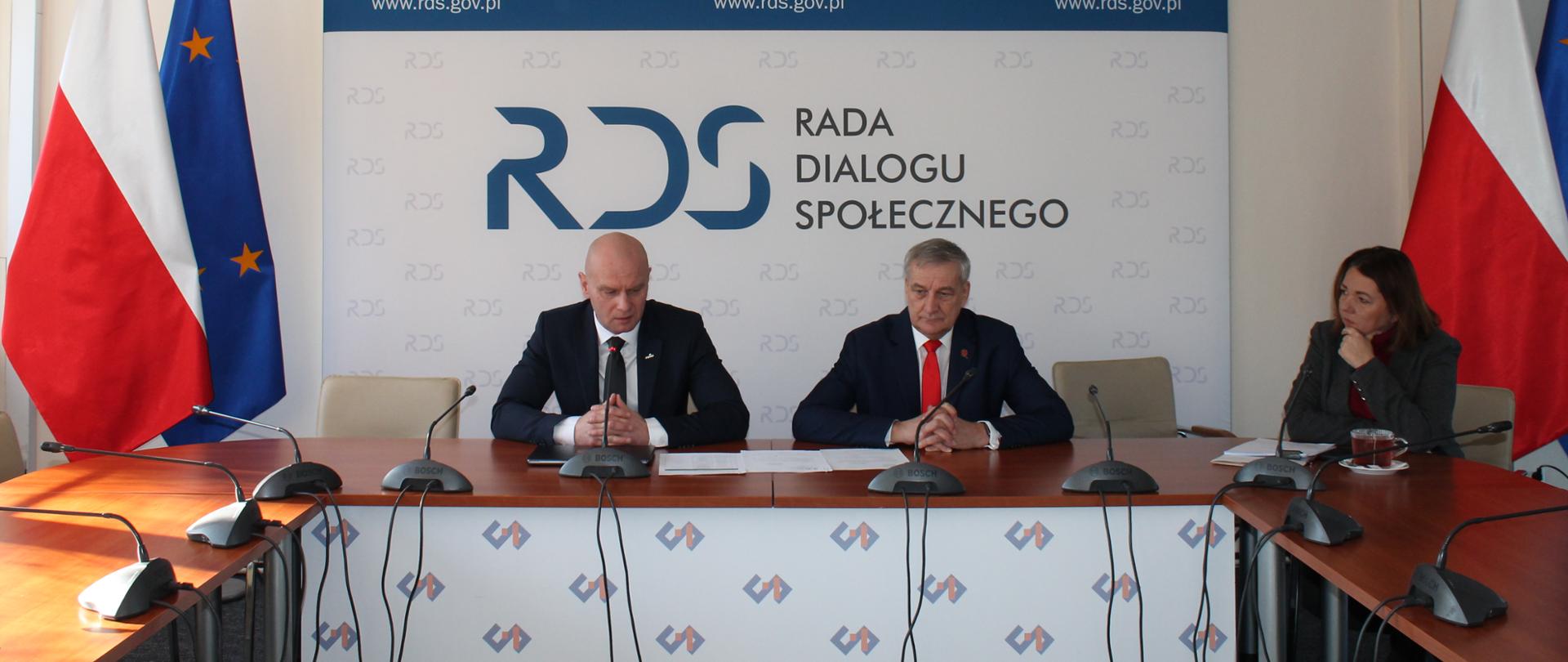 Na zdjęciu widnieje stół prezydialny za którym siedzą od lewej strony: Przewodniczący zespołu ds. usług społecznych – Bartłomiej Mickiewicz, Wiesław Szczepański – Sekretarza Stanu w Ministerstwie Spraw Wewnętrznych i Administracji, Izabela Ziętka – Podsekretarz Stanu w Ministerstwie Edukacji Narodowej. W tle siedzących znajduje się baner z napisem Rada Dialogu Społecznego a nad nim wisi godło Polski. Po obu stronach stołu flagi Polski i UE.