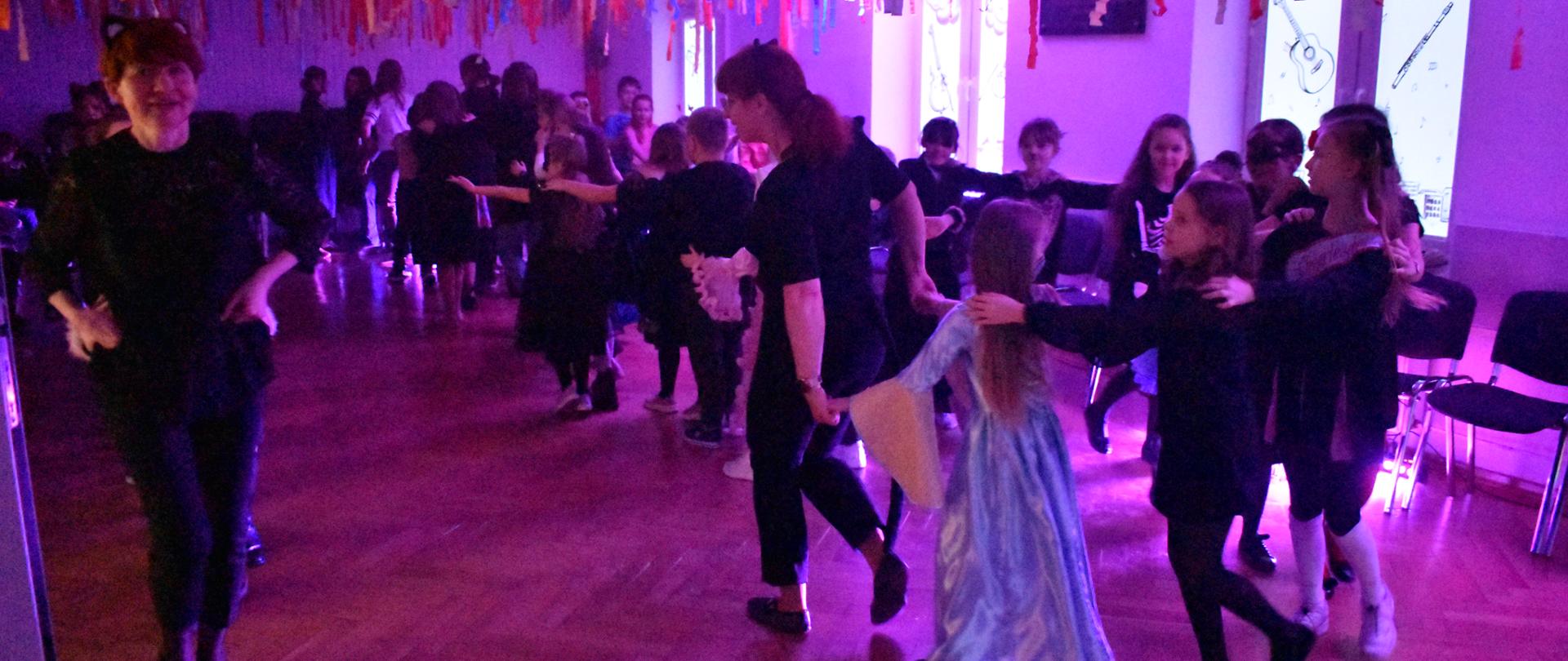 Zdjęcie przedstawia aulę PSM, w której odbywa się zabawa karnawałowa. Nauczyciele wraz z uczniami prowadzą tzw. „pociąg”. Kolorystyka zdjęcia jest różowo-fioletowo-czarna. 