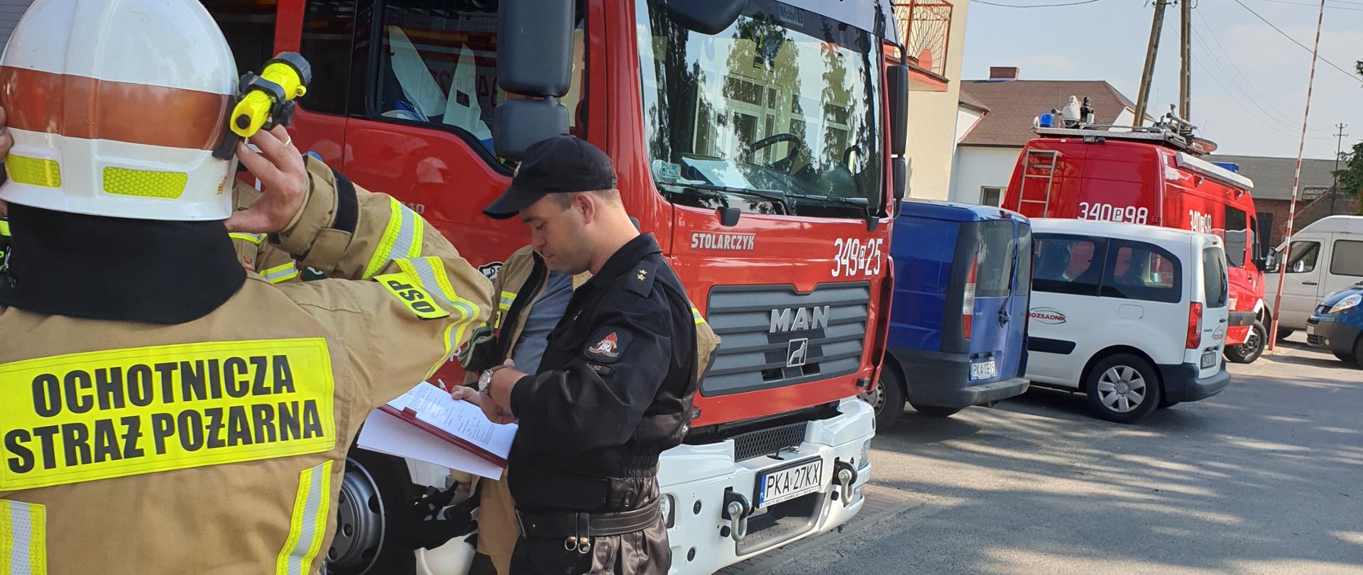 Na zdjęciu widać remizę OSP Rychnów, a przed nią samochód gaśniczy w kolorze czerwonym. Samochód posiada sygnalizację ostrzegawczą. Zderzak jest w kolorze białym. W środku zdjęcia widać funkcjonariusza Wydziału Operacyjno-Szkoleniowego KM PSP Kalisz. Po lewej stronie druhowie OSP ubrani w ubranie specjalne w kolorze piaskowym. Po prawej stronie zaparkowane samochody prywatne druhów. Osoby stoją na asfalcie w kolorze szarym.