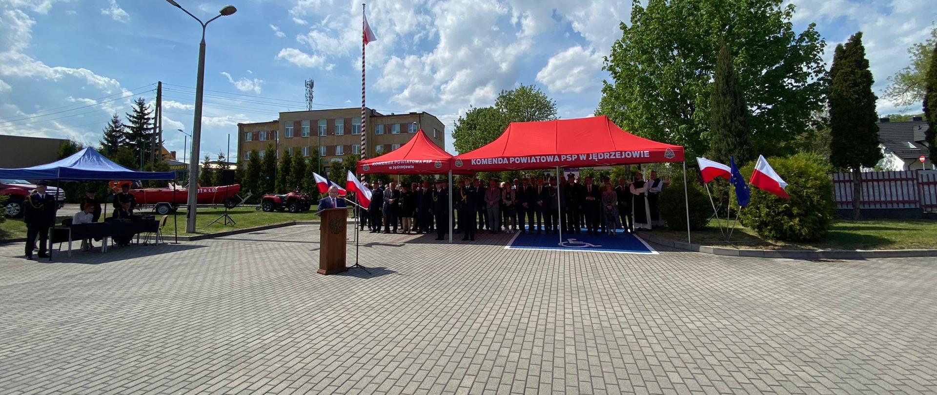 Zdjęcie przedstawia wicemarszałka województwa świętokrzyskiego Marka Bogusławskiego, który przemawia stojąc przy mównicy. Za jego plecami trybuna honorowa zaproszonych gości oraz pojazdy pożarnicze.