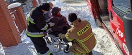 Po prawej stronie samochód straży pożarnej, po lewej stronie strażacy pomagają wsiąść do samochodu starszej kobiecie, która siedzi na wózku inwalidzkim