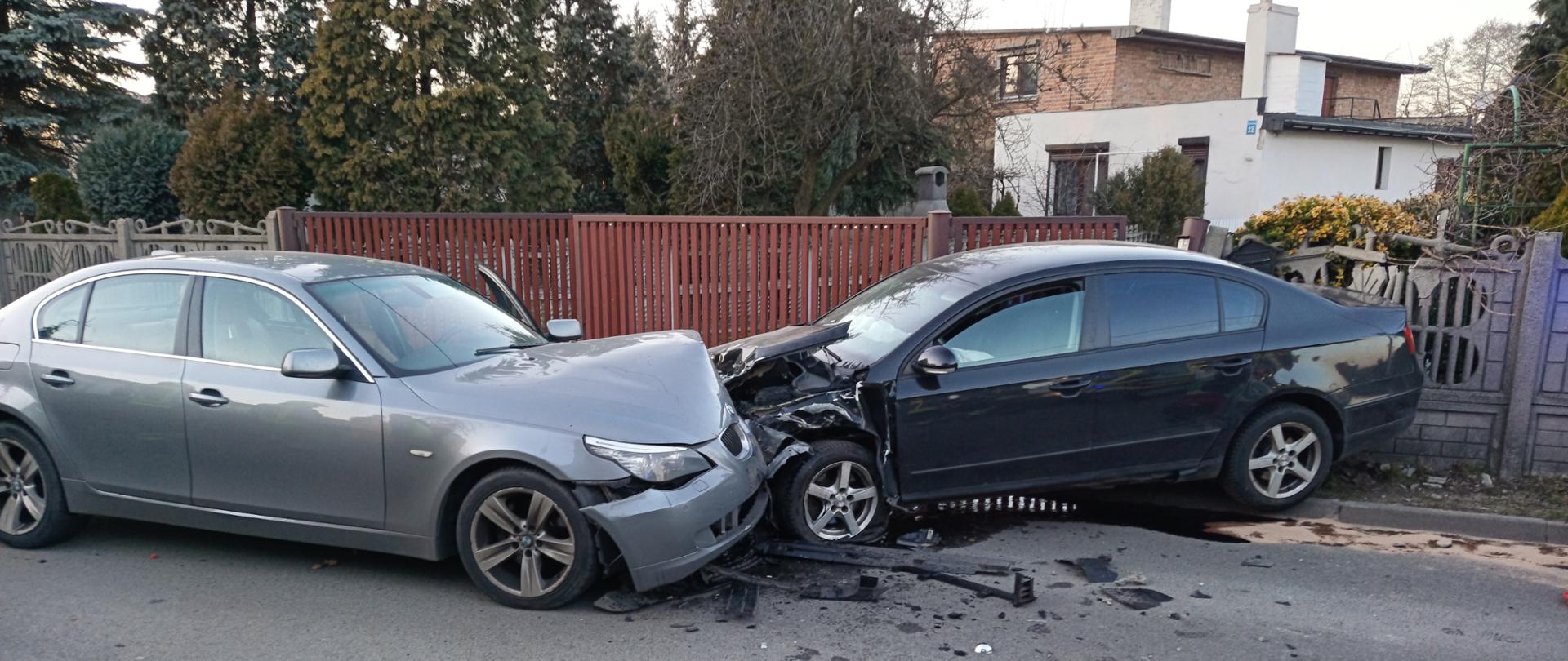 Na zdjęciu widać dwa pojazdy po zderzeniu. Auta uszkodzone. Jedno w kolorze jasnym to pojazd BMW, drugie w kolorze ciemnym VW. Pora dnia to dzień. W tle ogrodzenie posesji i brama.