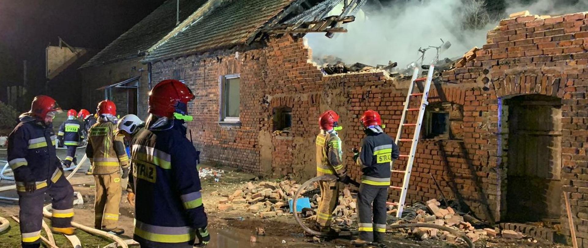 Zdjęcie przedstawia strażaków PSP podczas działań gaśniczych - pożar budynku gospodarczego w Kujakowicach Górnych. Na pierwszym planie strażacy PSP i OSP w czerwonych i białych hełmach na głowie i ubraniach specjalnych (piaskowy i czarny) - jeden trzyma odcinek strażacki. W tle widoczny ciąg budynków - w tym budynek gospodarczy - ze spalonym dachem części gospodarczej. Budynek zbudowany z cegły,