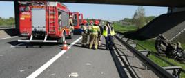 Zdjęcie przedstawia wypadek samochodu osobowego z ciężarowym na autostradę A4. Jest pora dzienna. Na zdjęciu widoczny jest średni samochód ratowniczo – gaśniczy GBARt 2,5/16 z JRG Bochnia o numerze 401 k 22 z otwartymi drzwiami kabiny. Za samochodem ustawione są biało czerwone stożki ostrzegawcze zabezpieczające miejsce zdarzenia. Na pasie awaryjnym stoi trzech strażaków oraz dwóch policjantów prowadzących czynności dochodzeniowe dotyczące wypadku. Za balustradą znajduje się zniszczony samochód osobowy biorący udział w wypadku. W tle widać fragment mostu, który znajduje się nad autostradą.
