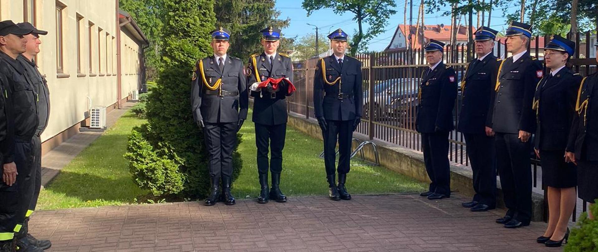Zdjęcie przedstawia strażaków zgromadzonych przy maszcie na którym podniesiona została flaga państwowa. Strażacy ubrani są w umundurowanie bojowe, a na głowach maja czerwone hełmy. Na pierwszym planie widać poczet flagowy podnoszący flagę do góry. Natomiast w tle widoczny jest Komendant Powiatowy w mundurze wyjściowym. W tle widać wysokie iglaste drzewa.