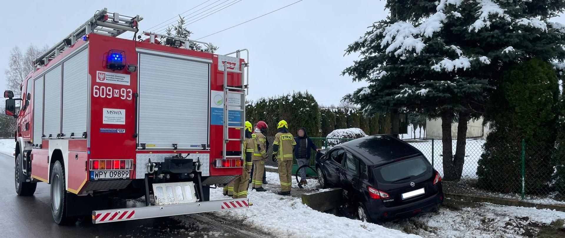 Zdjęcie przedstawia miejsce zdarzenia drogowego w zimowej scenerii. Na pierwszym planie po lewej stronie znajduje się tył czerwonego wozu strażackiego z numerem "609 93" i tablicą rejestracyjną "WPY 09899". Obok wozu, w rowie, widoczny jest czarny samochód osobowy, który zjechał z drogi i utknął w śniegu oraz błocie, zawisł na betonowym przepuście. W pobliżu czarnego samochodu stoi kilku strażaków w odblaskowych mundurach i hełmach, oceniających sytuację lub prowadzących działania ratownicze. Na ziemi wokół pojazdu widać biały śnieg i błoto. Po prawej stronie zdjęcia rośnie duże, iglaste drzewo pokryte śniegiem. W tle widoczny jest płot i fragmenty innych drzew oraz budynków. Niebo jest szare i zachmurzone, co wskazuje na typowy zimowy dzień w Polsce. Całość obrazu sugeruje interwencję straży pożarnej w związku z wypadkiem samochodowym w trudnych warunkach pogodowych.