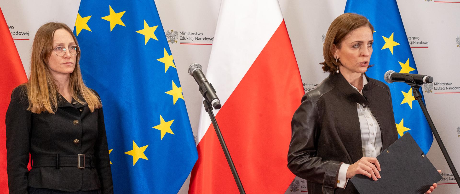 Wiceminister Joanna Mucha stoi przy mikrofonie i mówi, po lewej stronie stoi kobieta w ciemnej garsonce. W tle flagi biało-czerwone i niebieskie ze złotymi gwiazdkami. 