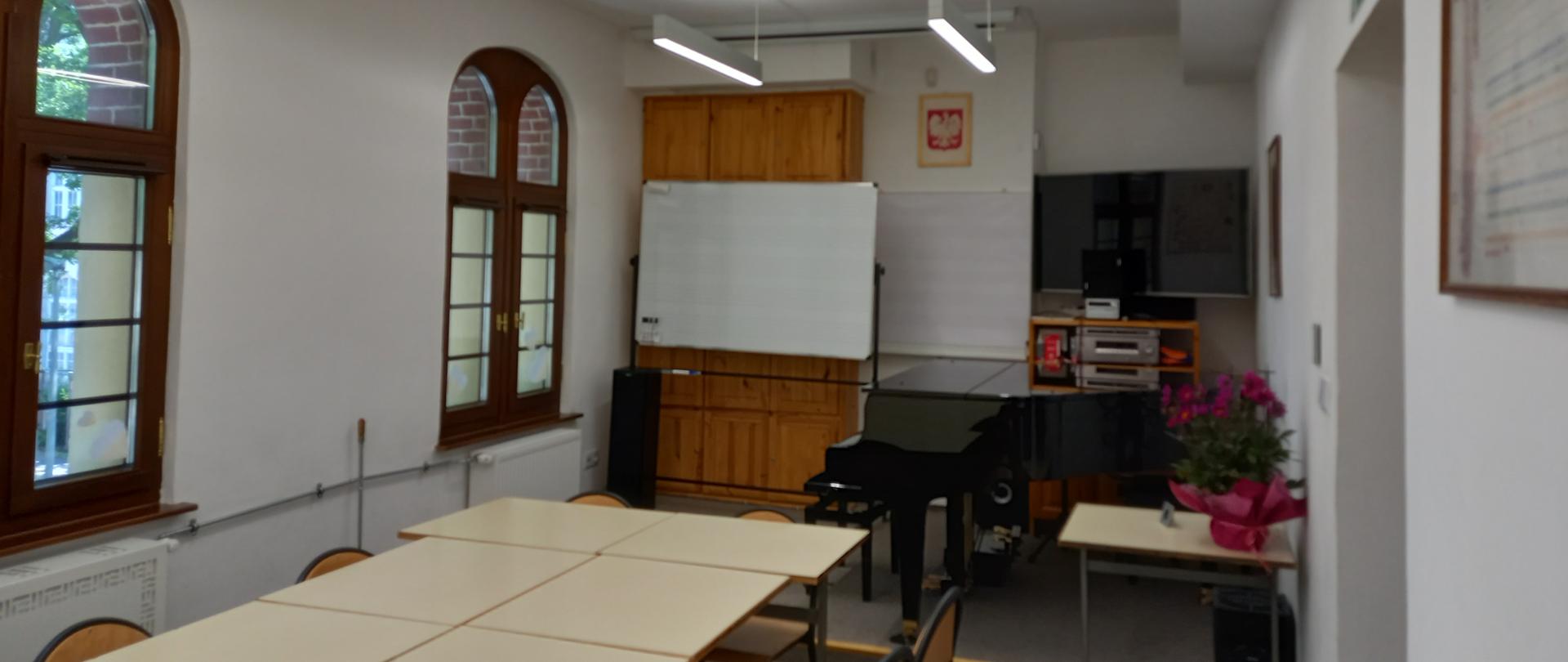 Zdjęcie przedstawiające salę w szkole muzycznej, białe ściany, po lewej stronie dwa łukowe okna, po prawej stronie drzwi, w głębi mała scena, na której stoi tablica szkolna po lewej stronie i po prawej stronie fortepian; nad fortepianem umieszczony jest telewizor, przed fortepianem kwiaty; środkiem klasy ustawiono stoły i krzesła