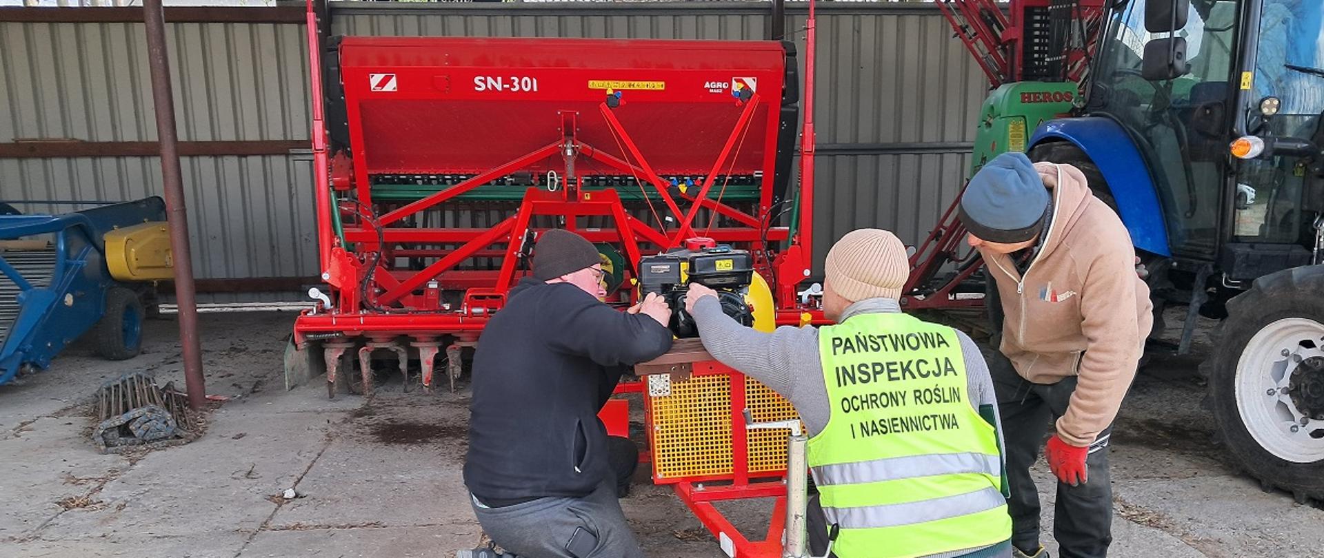 Trzech mężczyzn pochylonych przy urządzeniu rolniczym. Jeden z nich ma kamizelkę z logo PIORiN. Widoczny siewnik w kolorze czerwonym oraz niebieski ciągnik. 
