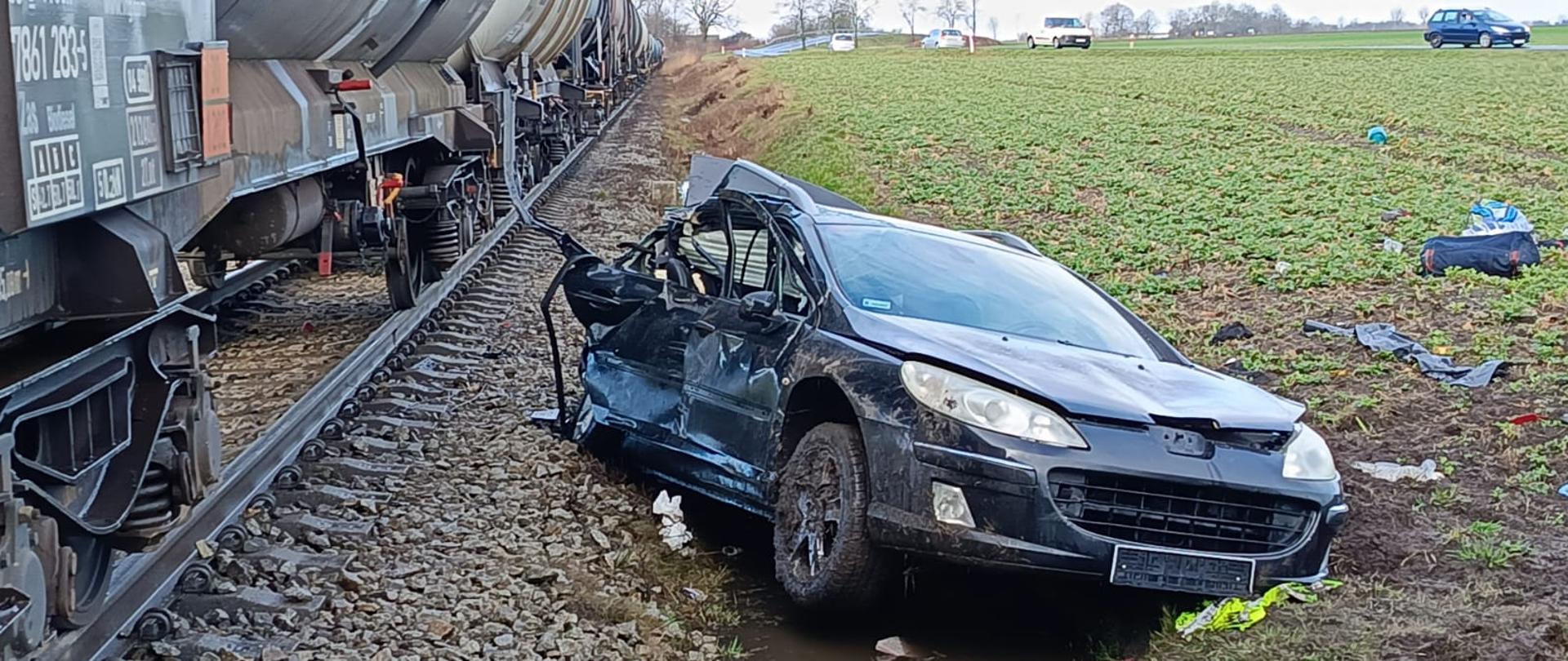 Tory kolejowe, na nich cysterny pociągu o kolorze brązowym. Na kamiennym nasypie rozbite czarne auto osobowe. 