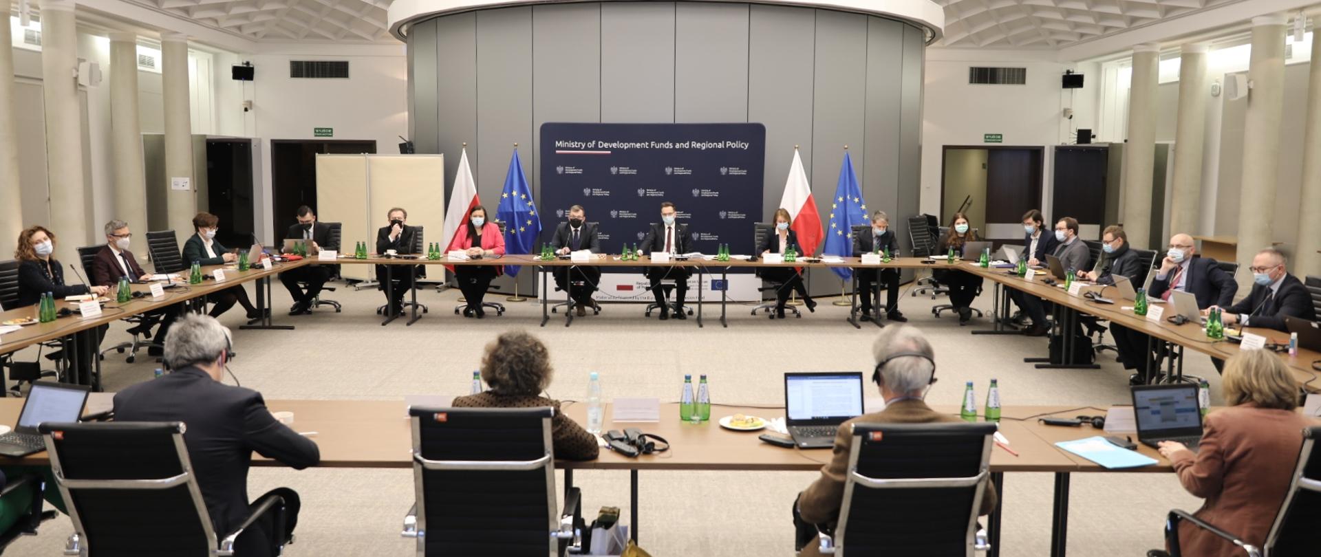 Na zdjęciu kilkanaście osób; siedzą za stołami ustawionymi na planie kwadratu, na stołach mikrofony, dokumenty i komputery. Na drugim planie ścianka z logotypami Ministerstwa Funduszy i Polityki Regionalnej oraz flagi Polski i Unii Europejskiej..