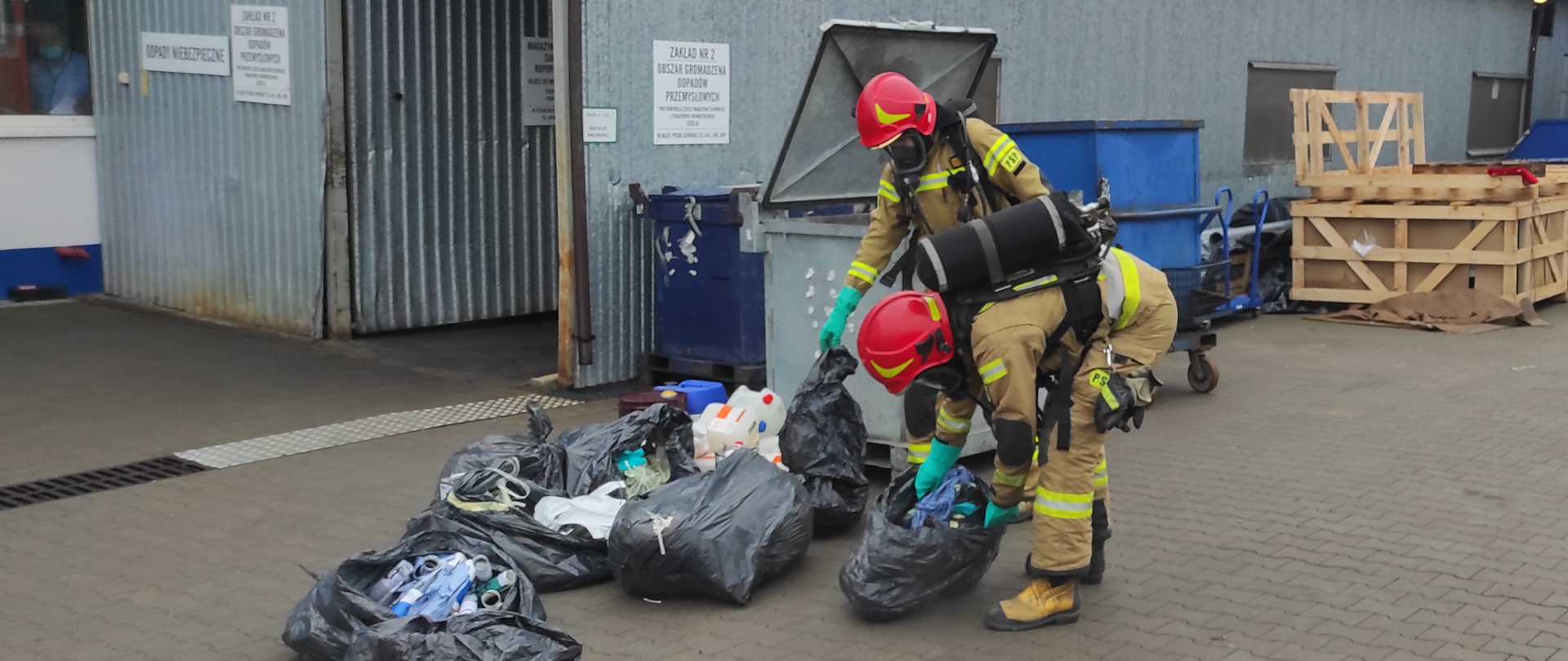 Na zdjęciu widzimy dwóch strażaków pochylonych nad ciemnymi workami na śmieci. Strażacy ubrani są w ubrania specjalne koloru piaskowego, na głowie czerwone hełmy, na twarzy maski ochrony dróg oddechowych , na plecach czarne butle z powietrzem. W tle widzimy kontener na śmieci oraz część hali koloru niebieskiego.
