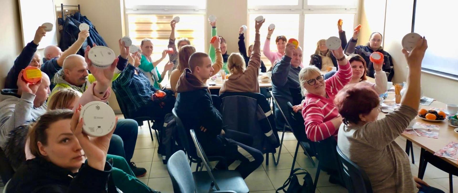 Osoby, uczestnicy spotkania trzymający w uniesionych rękach czujki dymu. 