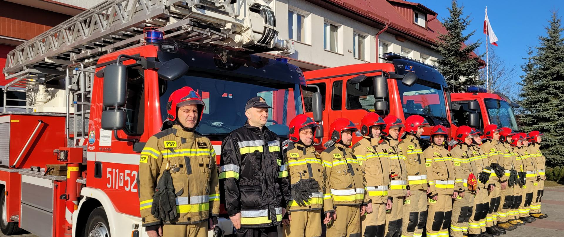 Strażacy w ubraniach specjalnych podczas uroczystego apelu na tle samochodów pożarniczych w dalszym planie budynek i maszt z flagą