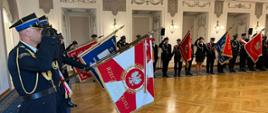 W reprezentacyjnej sali stoi grupa osób w galowych mundurach, trzymających sztandary i flagi. Uczestnicy uroczystości ustawieni są w szeregu, a część z nich oddaje honor podczas oficjalnego wydarzenia.