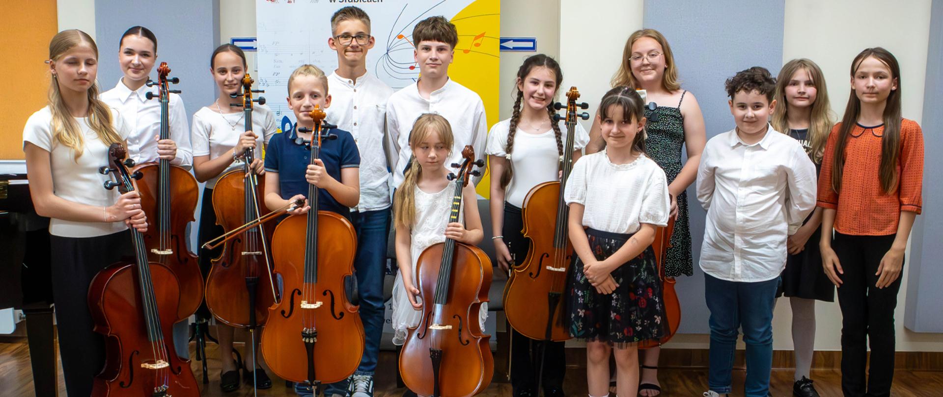 Na zdjęciu kilkanaścioro dzieci stoi w auli szkolnej, część z nich trzyma w rękach wiolonczele.