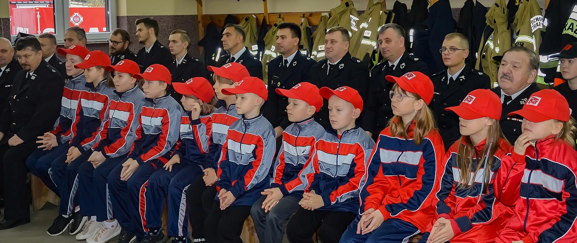 uczestnicy uroczystości wręczenia promes dla młodzieżowych drużyn pożarniczych wśród nich dzieci z młodzieżowej drużyny pożarniczej, strażacy druhowie, siedzą na ławkach w pomieszczeniu za nimi na wieszakach białe hełmy strażackiego oraz ubrania specjalne