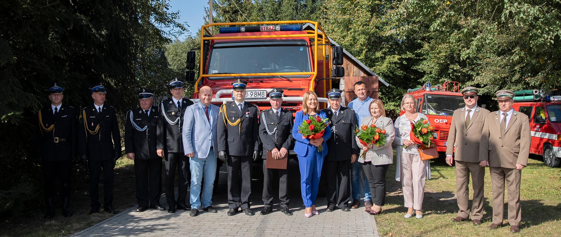 Zdjęcie przedstawia zaproszonych na uroczystość przekazania pojazdu gości ustawionych w rzędzie na tle pojazdu pożarniczego