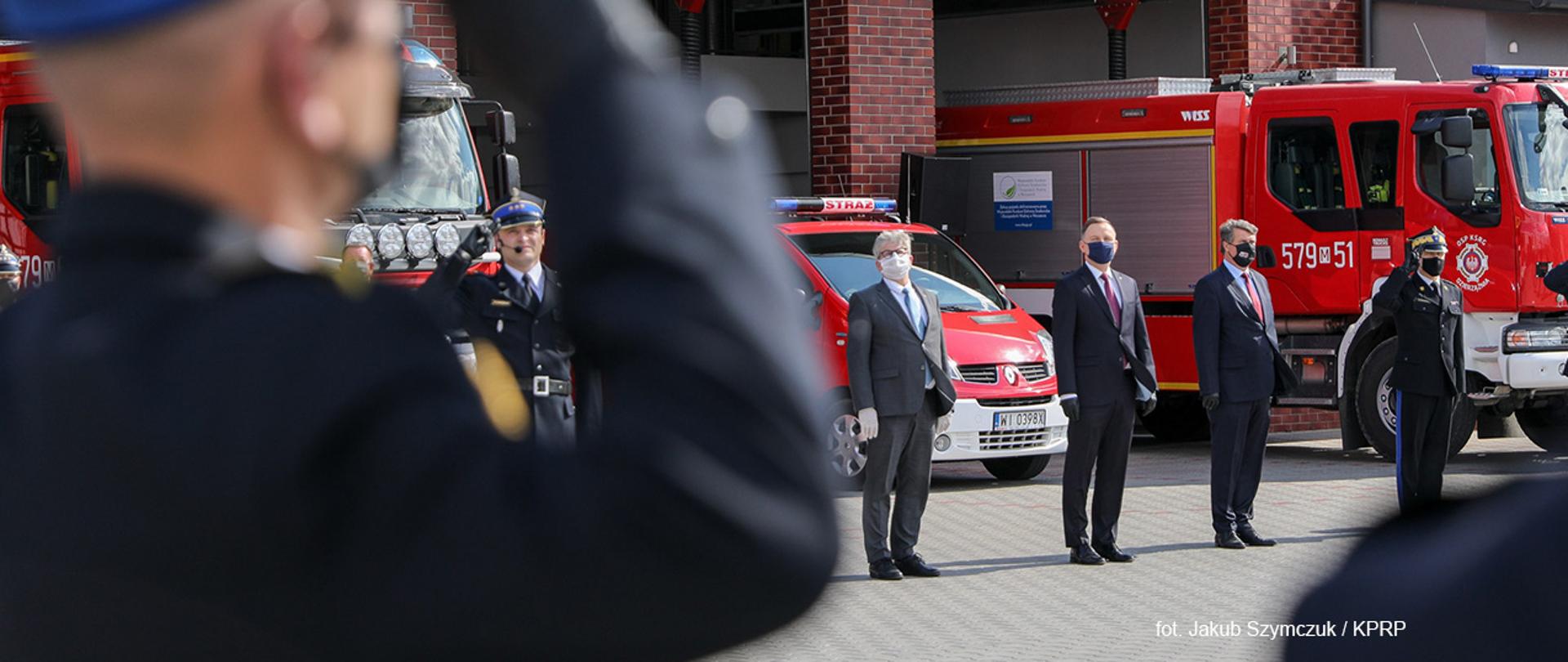 Uroczystość wręczenia promes na zakup samochodów dla OSP z Mazowsza, na zdjęciu m.in Prezydent RP, wiceminister MSWiA Maciej Wąsik. Na pierwszym planie widać nieostrego salutującego strażaka, w tle stojące samochody strażackie.