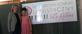 Na tle dużego napisu informaującego o konkursie stoją nauczycielka i jej uczennica. Uśmiechają się w stronę obiektywu.