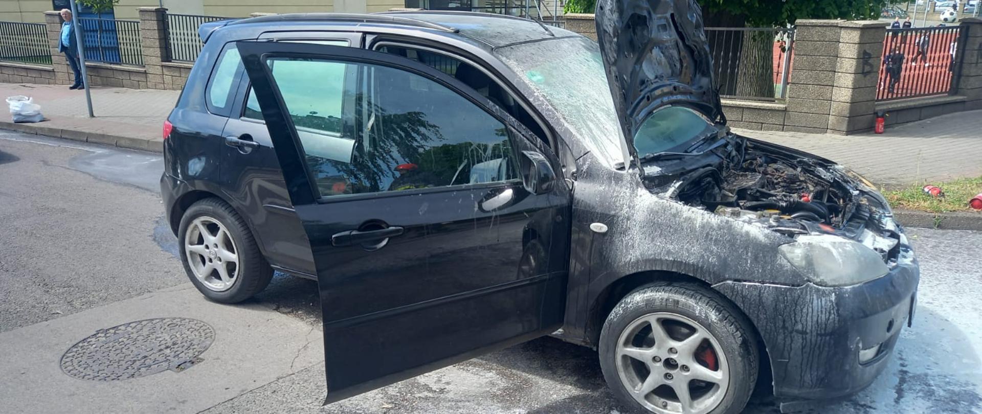 Zdjęcie przedstawia niebieski samochód osobowy z podniesioną maską po pożarze silnika. W tle znajdują się budynki oraz drzewa.