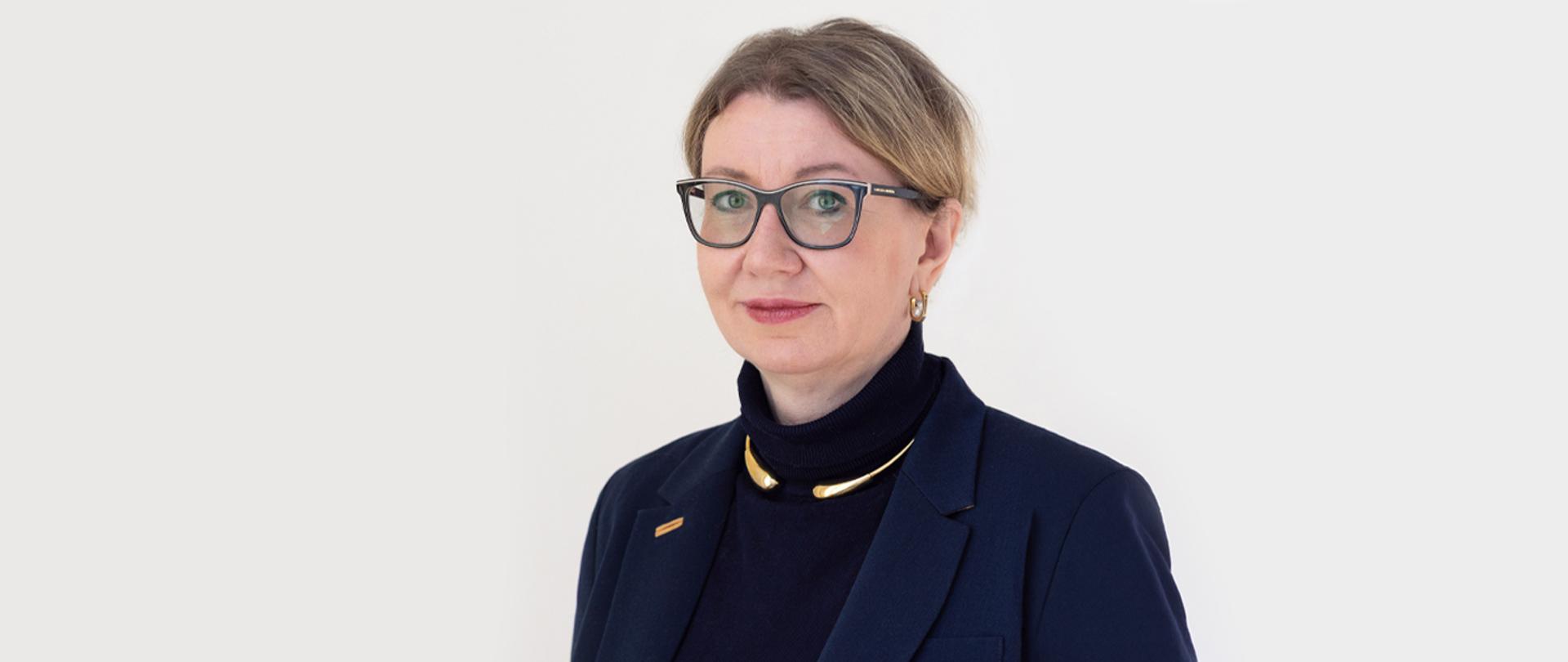 A middle-aged woman with glasses, formally dressed.