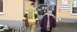 Zdjęcie zrobione na tle przychodni NZOZ w Lubomierzu. Strażak eskortuje starszą osobę w drodze do punktu szczepienia przeciw Covid-19. 