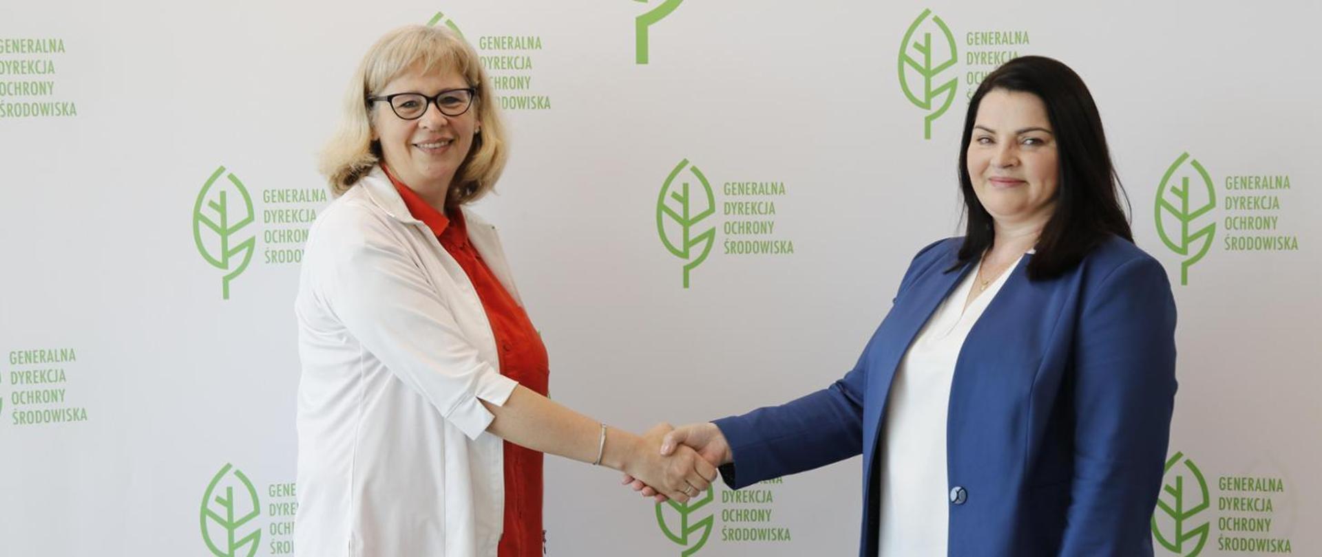 Na tle białej ściany z logotypem Generalnej Dyrekcji Ochrony Środowiska (zielony liść) stoją dwie uśmiechnięte kobiety. Ściskają sobie dłonie.