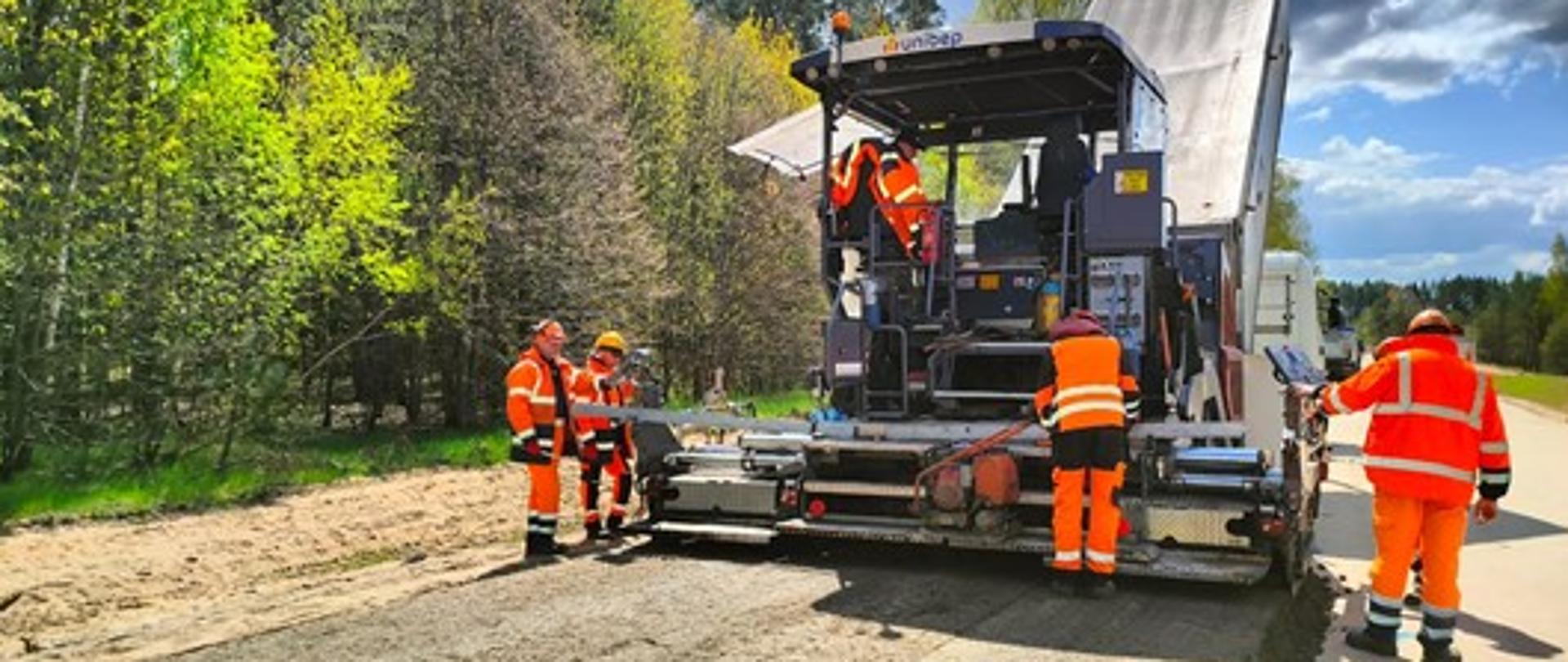 Czterech pracowników drogowych rozkłada mieszankę mineralno- cementową z asfaltem. Operator siedzi na maszynie do rozlewania mieszanki. Zdjęcie jasne zrobione przy słonecznej pogodzie,