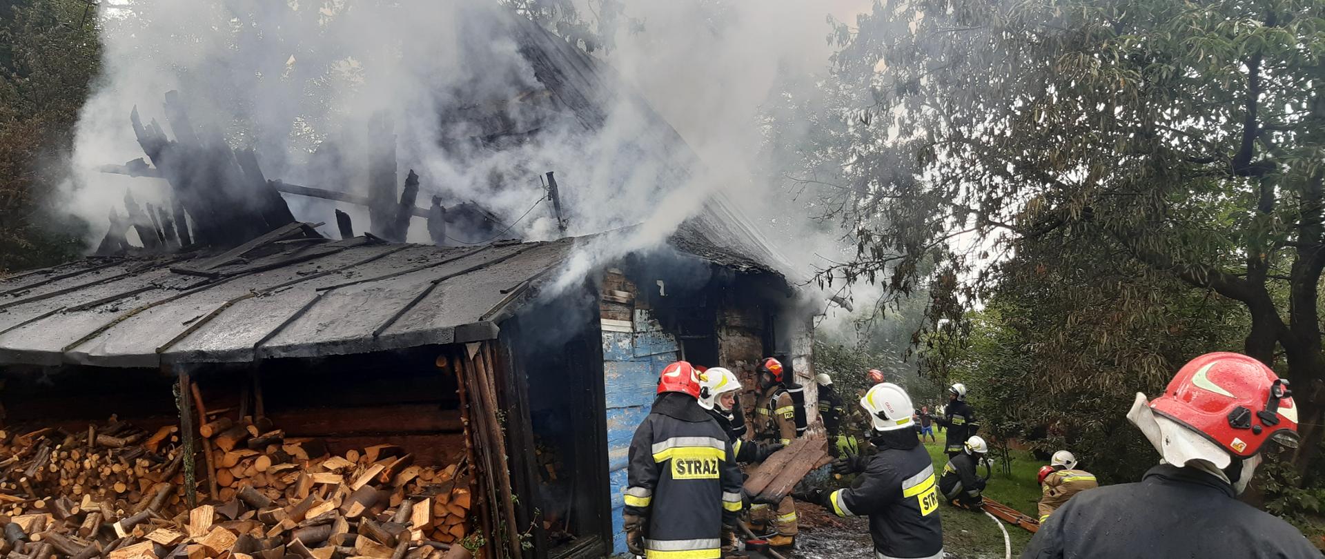 Na zdjęciu budynek drewniany częściowo ugaszony, spalona część dachu, obok strażacy zawodowi oraz ochotnicy dogaszają pożar 
